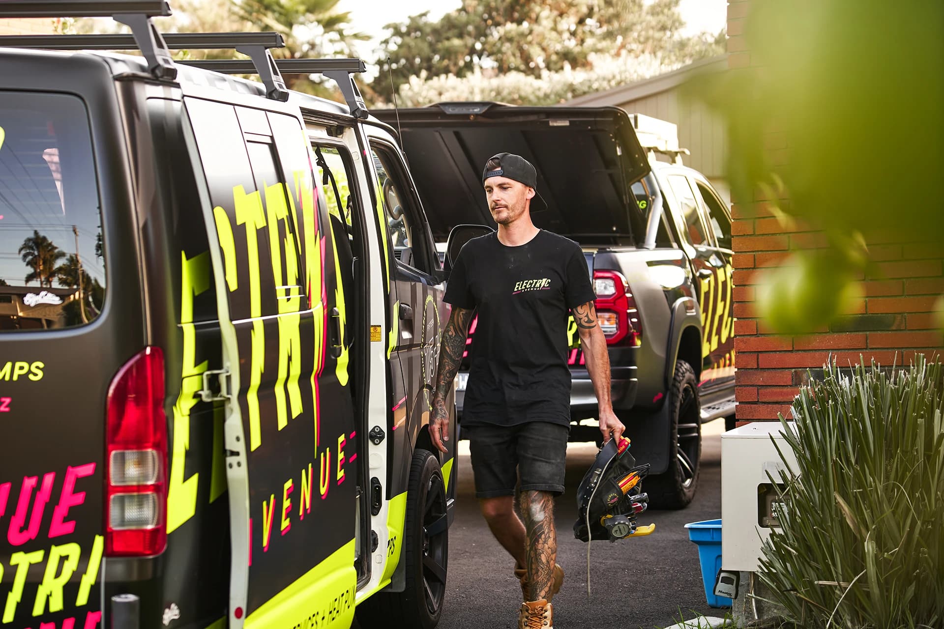 Electric Avenue electrician walking to a job with tools, branded work vans parked in driveway, Tauranga